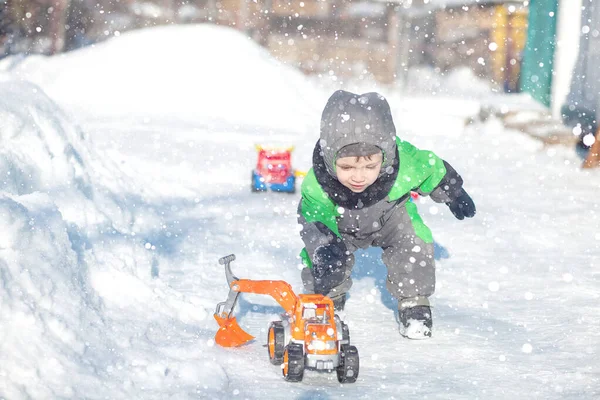 Ritratto di piccolo bambino carino seduto sulla neve e giocare con il suo giocattolo trattore giallo nel parco. Bambino che gioca all'aperto. Ragazzo felice con giocattolo da costruzione. Stile di vita concetto — Foto Stock