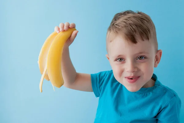 Маленький хлопчик тримає і їсть банан на синьому фоні, їжу — стокове фото