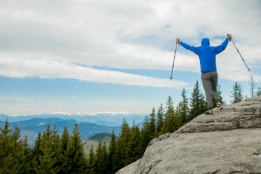 Dağlarda gökyüzüne karşı yükselen bir dağcı zaferi kutluyor, ellerini kaldırıyor.