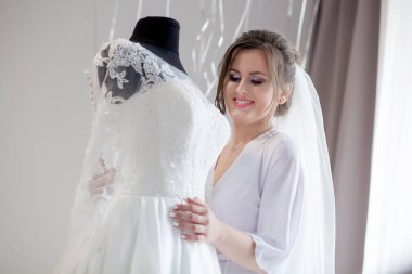 İpek bornozlu genç kadın gelinliğine dokunuyor ve pencerenin yanında dururken gülümsüyor. Düğün günü. Güzellik, moda. En mutlu düğün günü. Her anın tadını çıkar.