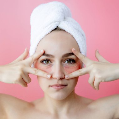 Kafasında beyaz havlu olan, göz altı kolajen bantlı pembe arka planda çıplak duran beyaz çilli kadının stüdyo fotoğrafı. Cilt bakımı, kozmetik ürün konsepti.