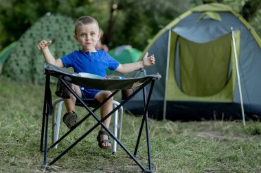 Küçük çocuk kamp alanında kahvaltı yapıyor. Güneş ışığı altında. Arka planda bir çadır var.
