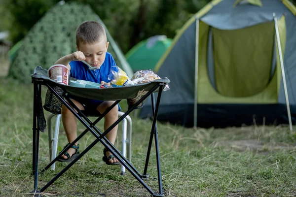 Küçük çocuk kamp alanında kahvaltı yapıyor. Güneş ışığı altında. Arka planda bir çadır var.