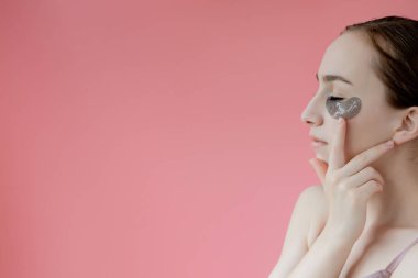 Baş portresi. Göz altı nemlendirici maskeli gülümseyen genç bir kadın. Kameraya bakıp cilt bakımının keyfini çıkarıyor..