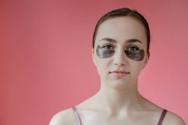 Baş portresi. Göz altı nemlendirici maskeli gülümseyen genç bir kadın. Kameraya bakıp cilt bakımının keyfini çıkarıyor..