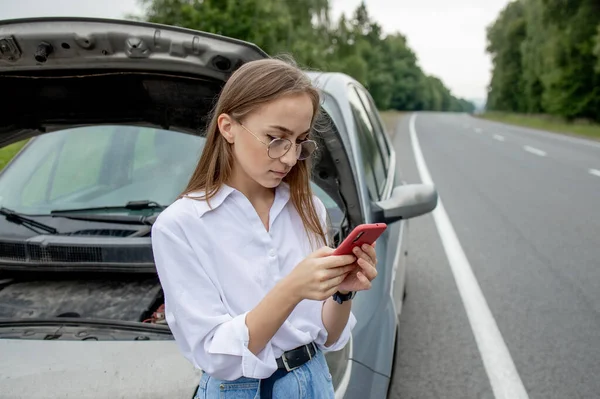 Young woman standing near broken down car with popped up hood having trouble with her vehicle. Waiting for help tow truck or technical support. A woman calls the service center.