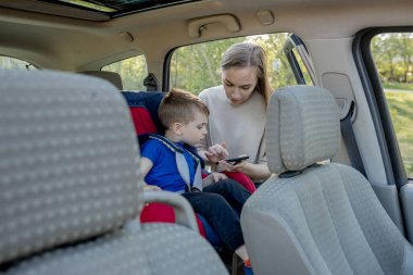 Anne, araba koltuğunda oturan küçük çocuğa telefonu verir. Çocuk taşımacılığının güvenliği