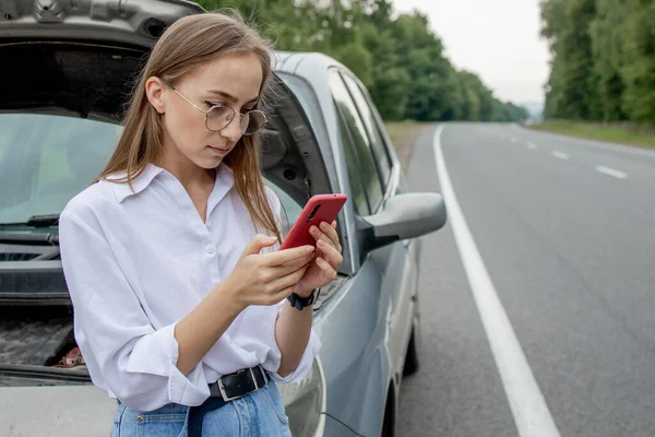 Young woman standing near broken down car with popped up hood having trouble with her vehicle. Waiting for help tow truck or technical support. A woman calls the service center.