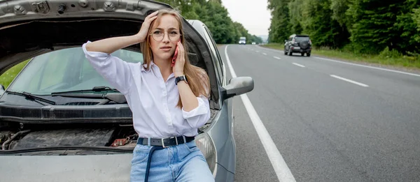 若い女性が壊れた車のボンネットを開けると、彼女の車に問題があった。心配している女性壊れた車の近くの電話で話している。自動車サービスのための壊れた車の呼び出しの近くに立つ女の子モデル — ストック写真