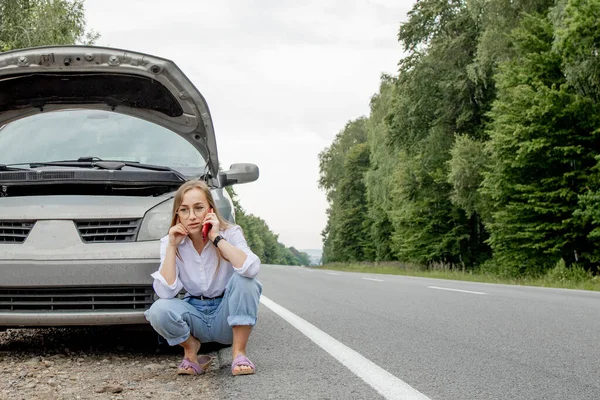 壊れた車の近くに立っている若い女性は 彼女の車に問題があるフードをポップアップしました トラックや技術サポートを牽引するのを待っています 女性がサービスセンターに電話して — ストック写真