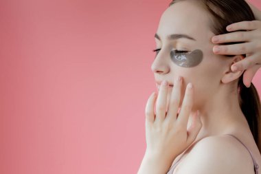 Baş portresi. Göz altı nemlendirici maskeli gülümseyen genç bir kadın. Kameraya bakıp cilt bakımının keyfini çıkarıyor..