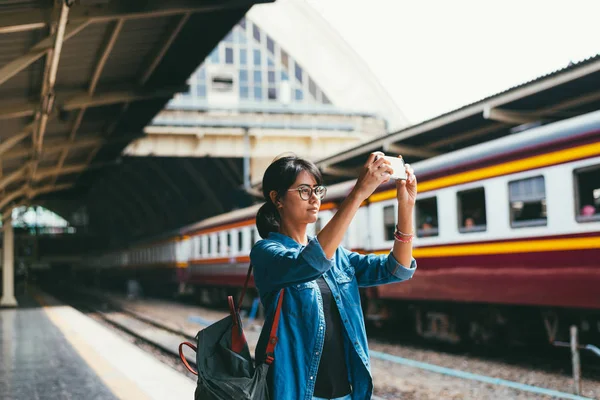 Asian woman traveler used smart phone take photos while wait and check train schedule on the platform of the railway station - travel and transportation concept