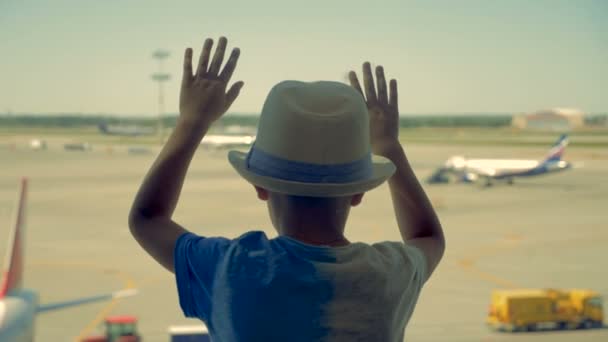 En pojke i en hatt ser på flygplan genom fönstret på flygplatsen. — Stockvideo