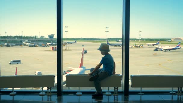 Een jongen kijkt vliegtuigen door een raam wachten op een vlucht op de luchthaven. — Stockvideo