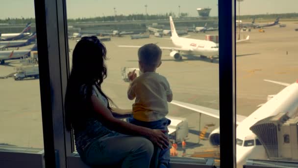 Mulher e rapaz olhem para a pista juntos no aeroporto, de perto. Uma família espera por um voo, enquanto observa aviões . — Vídeo de Stock