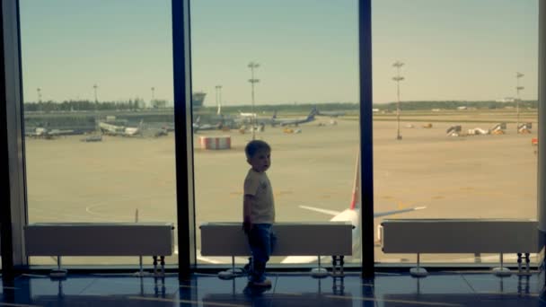 Un chico juega en la sala del aeropuerto. Un niño mira a la cámara mientras juega cerca de la ventana en el aeropuerto . — Vídeos de Stock