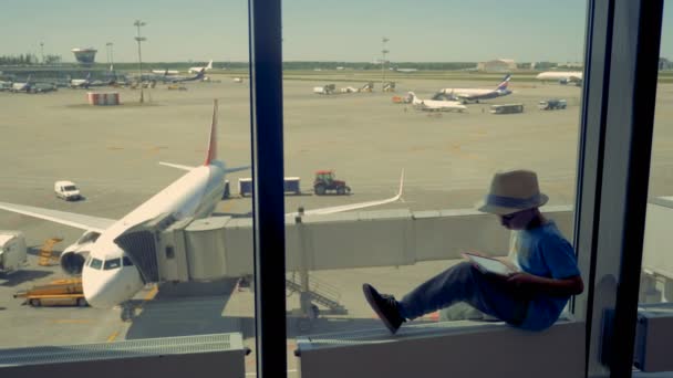 Ein Junge spielt in der Lounge mit einem Tablet. Kind spielt mit Tablet, während sich ein Flugzeug auf dem Rollfeld auf den Flug vorbereitet. — Stockvideo