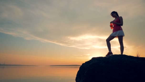 Une coureuse boit de l'eau, au ralenti. Une femme se tient sur un rocher près de l'eau, buvant sur un fond de coucher de soleil . — Video