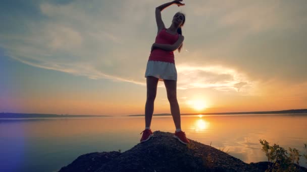 Une femme s'étend près du lac. Une fille fait des exercices dehors sur un fond de coucher de soleil . — Video