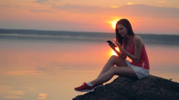 En kvinna ser på sin telefon på en solnedgång bakgrund. — Stockvideo