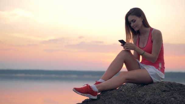 Chica de fitness se sienta cerca de un lago con un teléfono . — Vídeo de stock