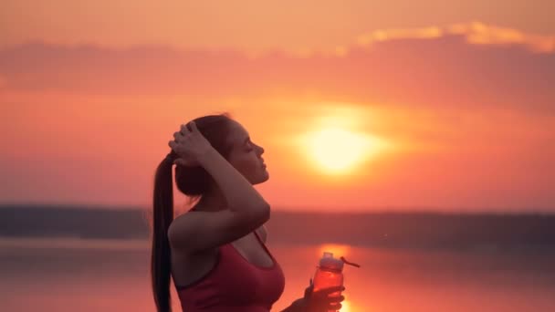 Pretty lady with a ponytail is standing near a lake and drinking fresh water from a bottle. — Stock Video