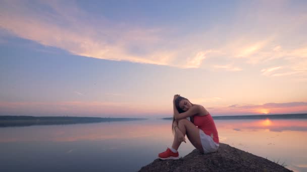Csinos fiatal nő békésen ül egy kövön, egy tó közelében — Stock videók