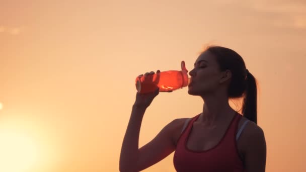 Гарна молода жінка п'є з апельсинової пляшки в променях сонця — стокове відео