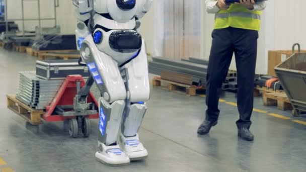 Un humano y un robot caminan juntos en un almacén, de cerca . — Vídeo de stock