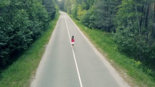 Vista aérea de un camino forestal y una joven delgada corriendo a través de él — Vídeo de stock
