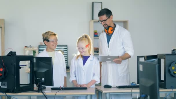 Un trabajador de laboratorio enseña a los niños a trabajar con una impresora 3D . — Vídeo de stock