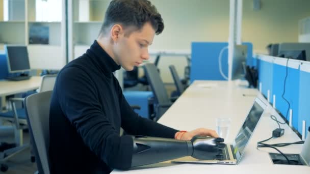 Un uomo si siede a un tavolo, lavorando con un computer portatile mentre indossa una mano protesica. 4K . — Video Stock