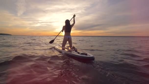 Paisaje acuático al atardecer con una señora en bikini paddleboarding en ella — Vídeo de stock
