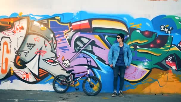 Stylish teenager cyclist stands near graffiti wall with his bike in a modern skatepark. — Stock Video