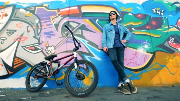 Hombre ciclista se apoya en una pared de graffiti con su bicicleta cerca de ella — Vídeos de Stock