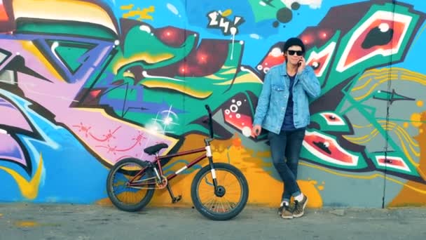 Graffiti zeď a mladý cyklista stojící proti ní a mluví o telefonu — Stock video