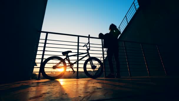 Erkek genç bir platform üzerinde dururken onun saçlı bisikletiyle günbatımında karıştırıyor — Stok video