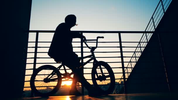 Hombre adolescente está sentado en su bicicleta en los rayos del sol poniente y quitándose el sombrero — Vídeos de Stock