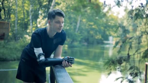 Un joven con un brazo biónico está parado cerca del puente del río. — Vídeo de stock