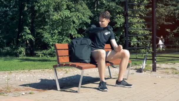 Подросток вытаскивает ноутбук из сумки протезной рукой, сидя на скамейке. — стоковое видео