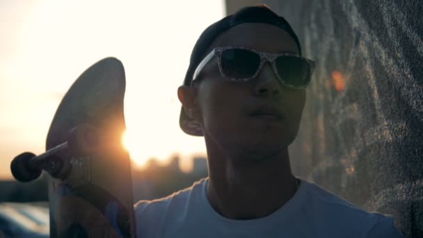 Retrato de un joven patinador con un monopatín sobre un fondo al atardecer . — Vídeo de stock