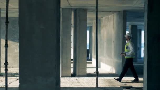 Ingénieur en vêtements de travail se promène le long du bâtiment en cours de construction . — Video