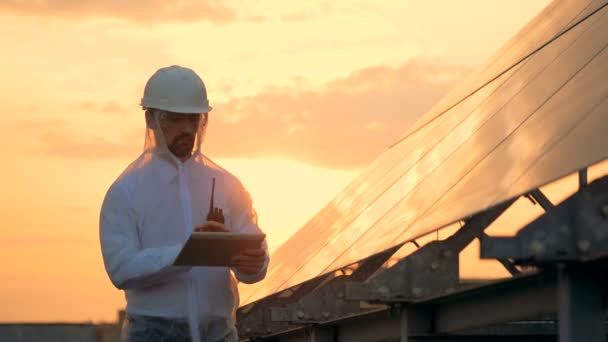 Sunset landscape with a male constructor operating a computer while standing beside a solar battery — Stock Video