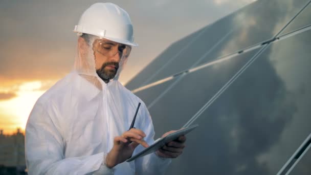 Tankeväckande solar arbetare står med en dator bredvid en solar installation i solnedgången. — Stockvideo