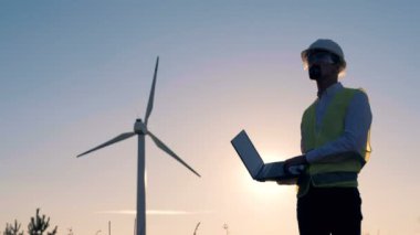 Erkek mühendis bir laptop tutar, üzerinde türleri ve elektrik yel değirmenleri denetler. Yenilenebilir electririty, yeşil enerji kavramı.