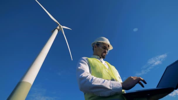 Ein Mann tippt auf einem Laptop in der Nähe einer elektrischen Windmühle, die auf einem Feld steht. Alternatives Energiekonzept. — Stockvideo