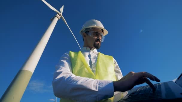 Een man werkt met een laptop in de buurt van een witte windturbine. Alternatieve energieconcept. — Stockvideo