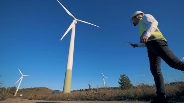 Ingenjören promenader i ett fält nära väderkvarnar, hålla en tablett. Alternativ energikoncept. — Stockvideo