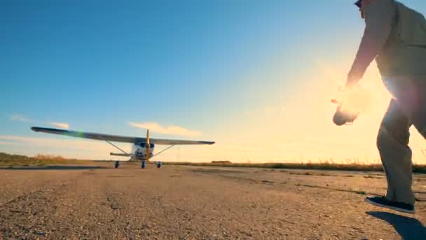 Pista iluminada por el sol con un aviador macho caminando hacia un avión y revisando su hélice — Vídeo de stock