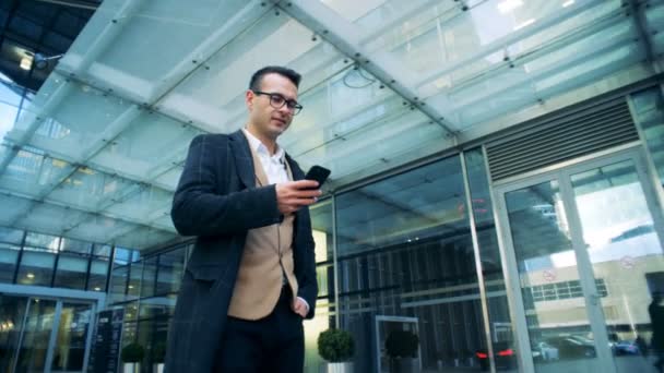 Smiling manager is walking next to an office center with a smartphone. Red epic cinema camera shot. — Stock Video